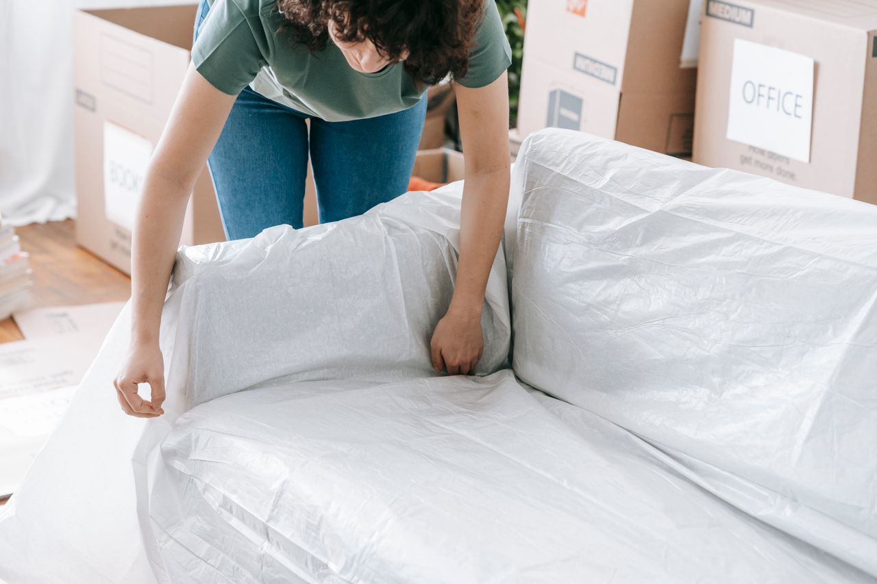 Une femme enveloppe soigneusement un canapé dans un matériau de protection tout en remplissant des cartons dans une pièce lumineuse.